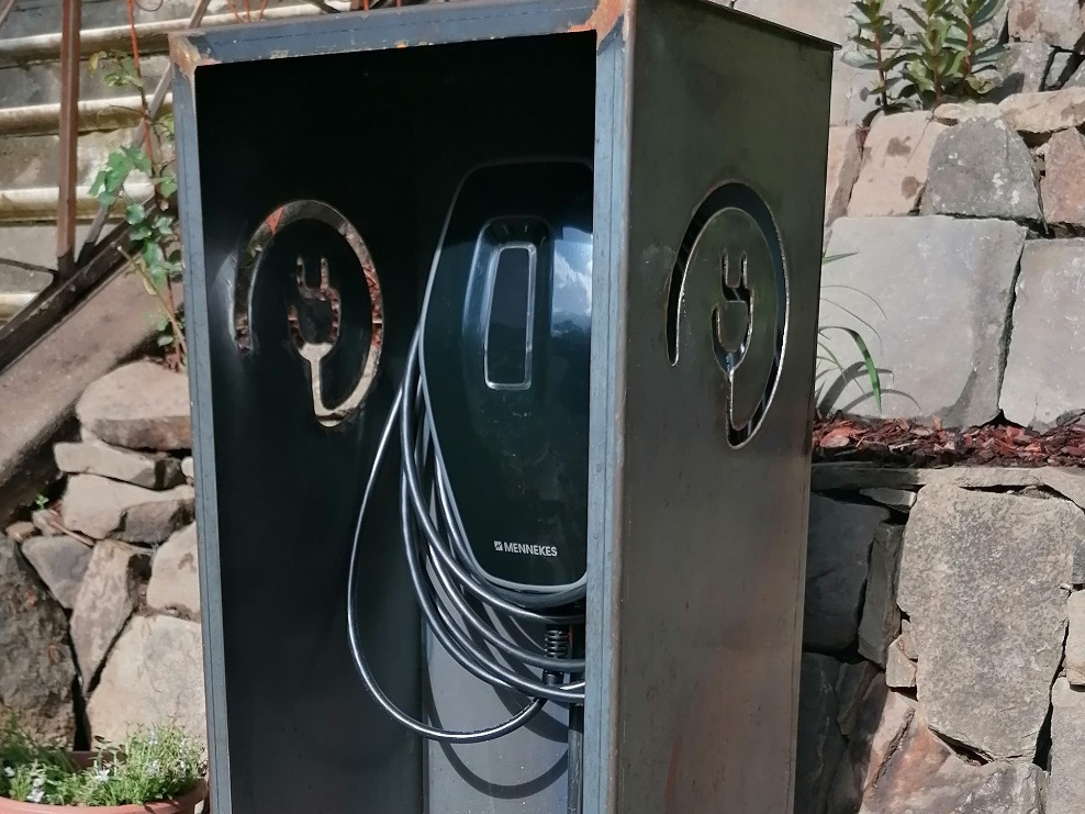 Geöffnete Wallbox-Verkleidung aus Cortenstahl mit sichtbarer Ladestation und Ladekabel an der Auffahrt.