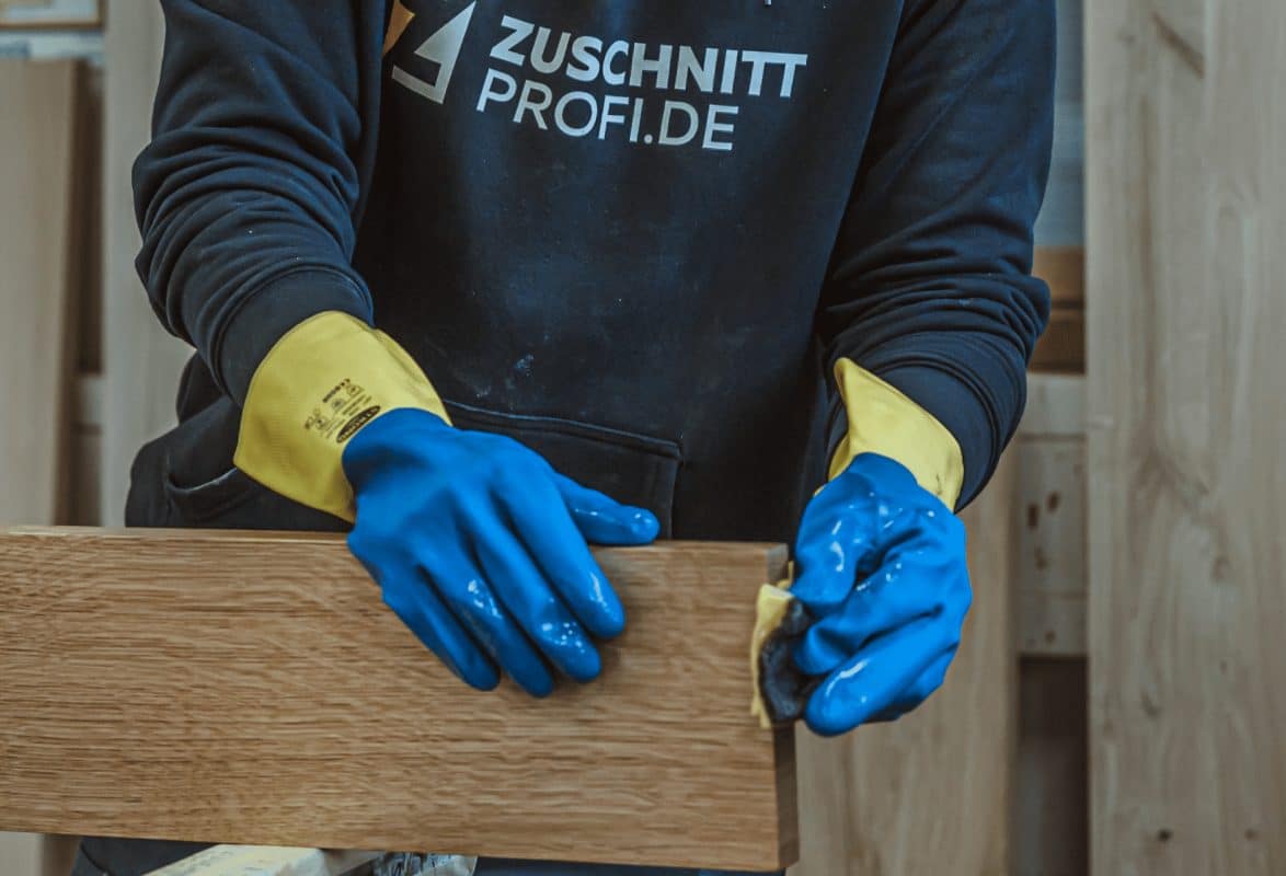 Ein Mitarbeiter von Zuschnittprofi.de arbeitet mit Handschuhen an einem Holzstück und verarbeitet die Oberfläche mit ruhiger Hand.