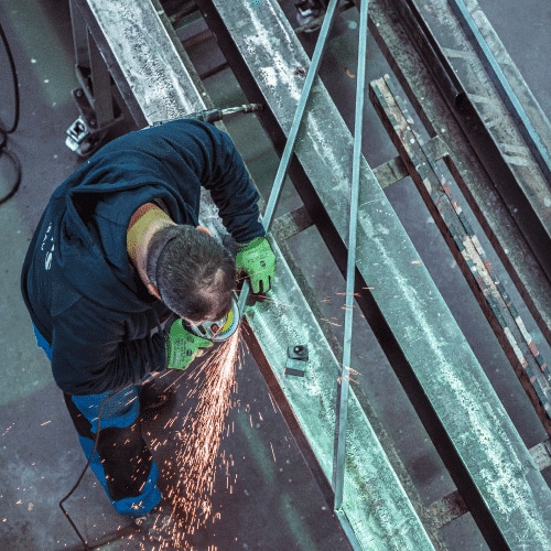 Ein Mitarbeiter von Zuschnittprofi.de arbeitet mit einem Winkelschleifer an großen Metallträgern und sorgt für saubere Schnittkanten.